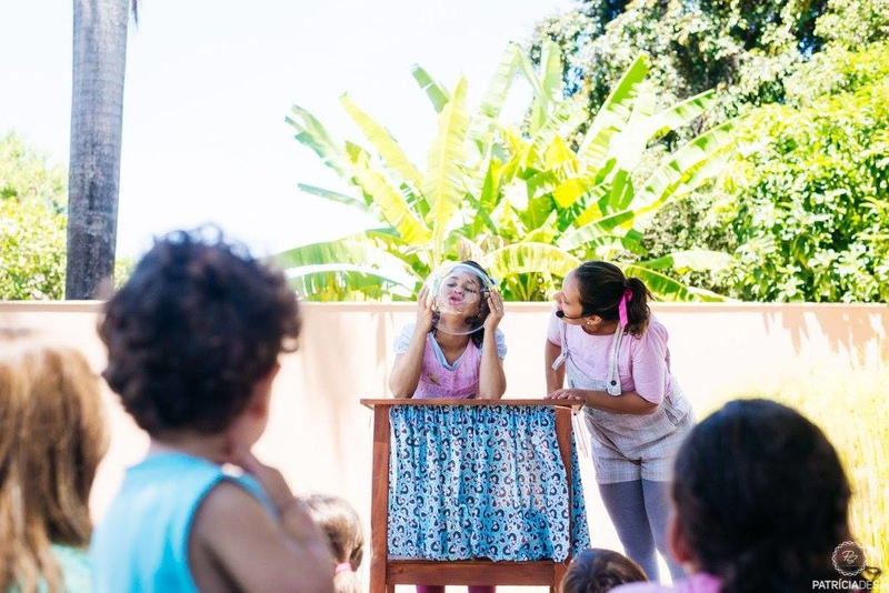 Em um quinta, crianças (de costas para a fotógrafa) assistem duas contadoras de histórias, vestidas de rosa e azul, sob uma mesa de madeira, segurando uma bacia de vidro. É a história do bonequinho doce. 