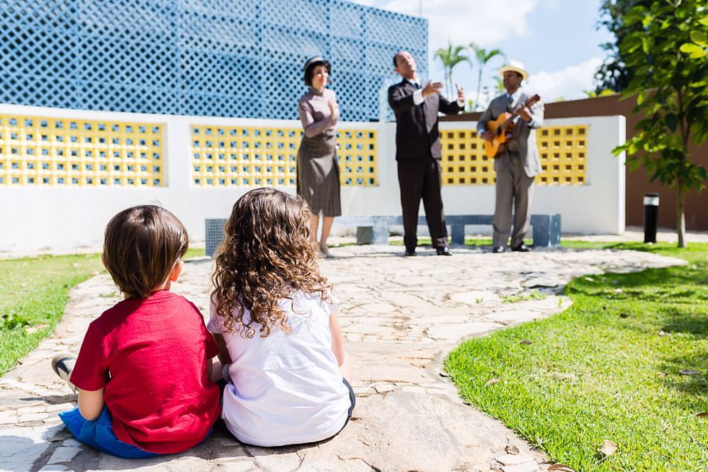 Foto colorida. Em um jardim, duas crianças observam 3 atores representando uma peça teatral.