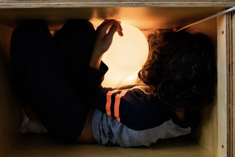um menino deitado dentro de um caixote de madeira brinca com uma bola de luz 