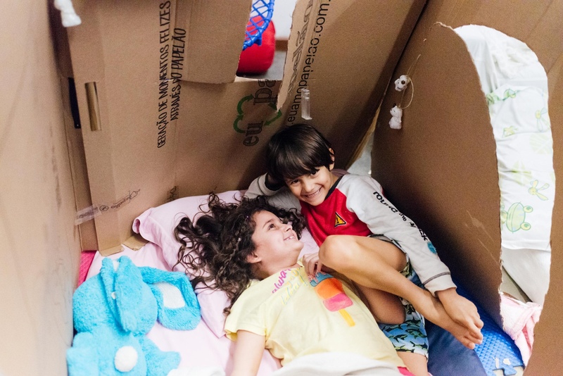 Duas crianças - um menino e uma menina - brincam de acampamento em uma casinha de papelão feita e decorada por eles