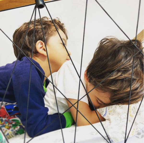 Foto de dois meninos irmãos de pele clara, cabelos castanhos, deitados em uma cama. O mais velho com a cabeça abaixada, recostada no travesseiro, e o mais novo o abraçando.