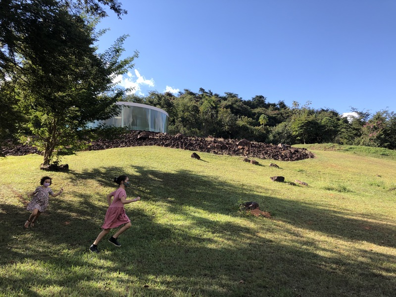 foto de duas meninas brancas, com cabelos castanhos presos, de vestido rosa e estampado, correndo em um gramado. ao fundo, a galeria Dougen Aitken. num dia de céu azul.