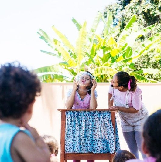 Em um quinta, crianças (de costas para a fotógrafa) assistem duas contadoras de histórias, vestidas de rosa e azul, sob uma mesa de madeira, segurando uma bacia de vidro. É a história do bonequinho doce.