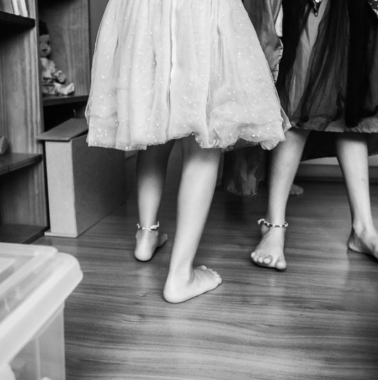 Duas meninas brincam em uma sala vestidas com saias de tule. O corte da imagem é na altura dos joelhos.