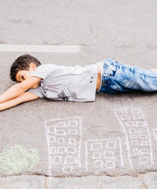 Menino deitado sob o asfalto brinca de voar em cima de prédios desenhados com giz no chão