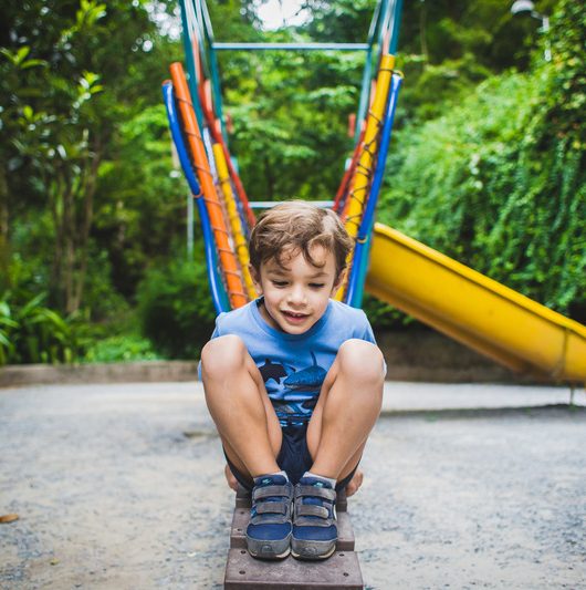 Foto colorida. Uma criança está sentada em uma rampa de madeira de um brinquedo em um parque. Ela sorri para o espectador.