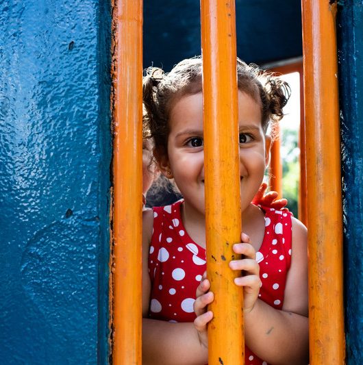 foto colorida. uma criança está atrás de um gradil laranja de um brinquedo. ela sorri para o espectador enquanto se esconde.