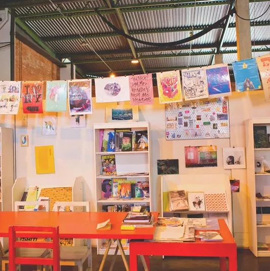 Ambiente interno de um centro cultural. Em primeiro plano, uma mesa vermelha com livros e gibis, ao fundo, estante com mais livros e uma parede colorida por cartazes com várias artes.